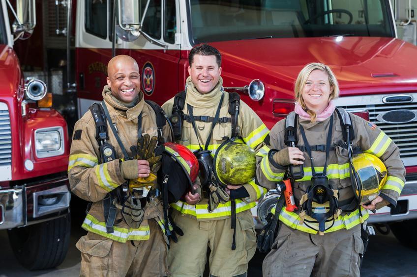 Image: three firefighters posing in front of two fire trucks